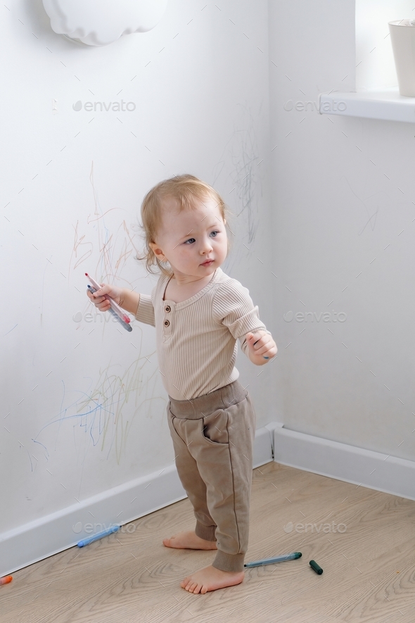 A cute toddler standing by the drawn white wall holding colored markers.