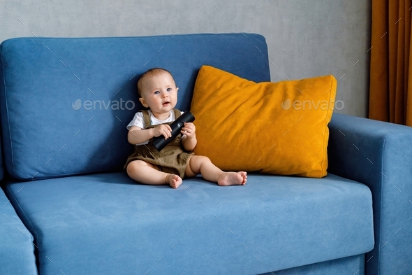 A cute baby sitting on a sofa, holding a remote control and watching cartoons on TV.