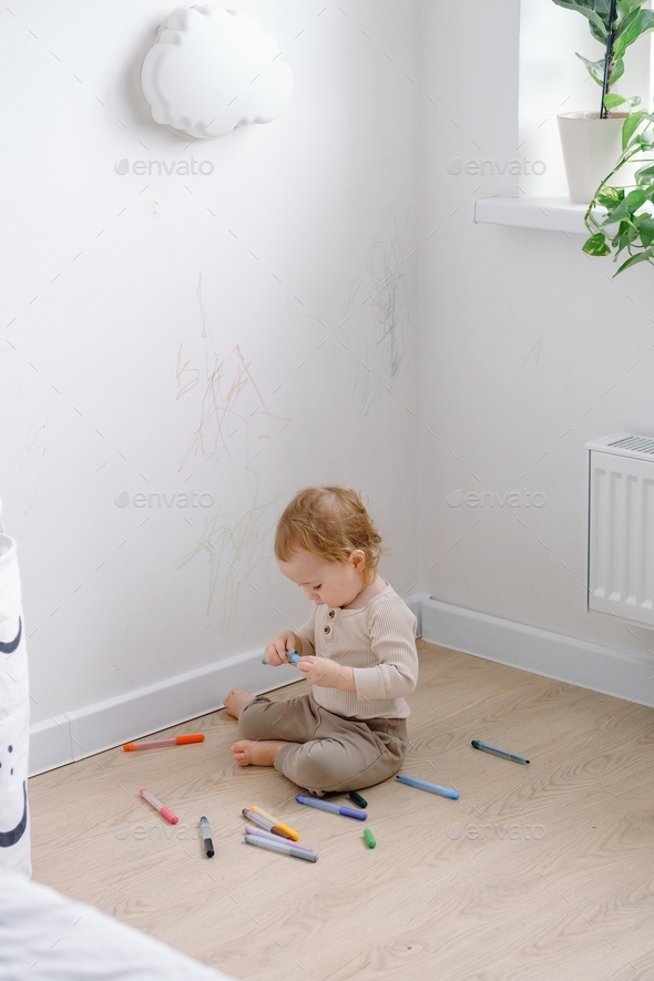 A toddler sitting by the white wall holding colored markers and drawing on the wall.