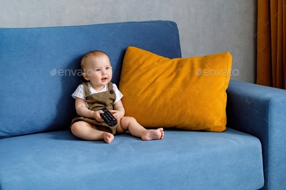 A cute baby sitting on a sofa, holding a remote control and watching cartoons on TV.