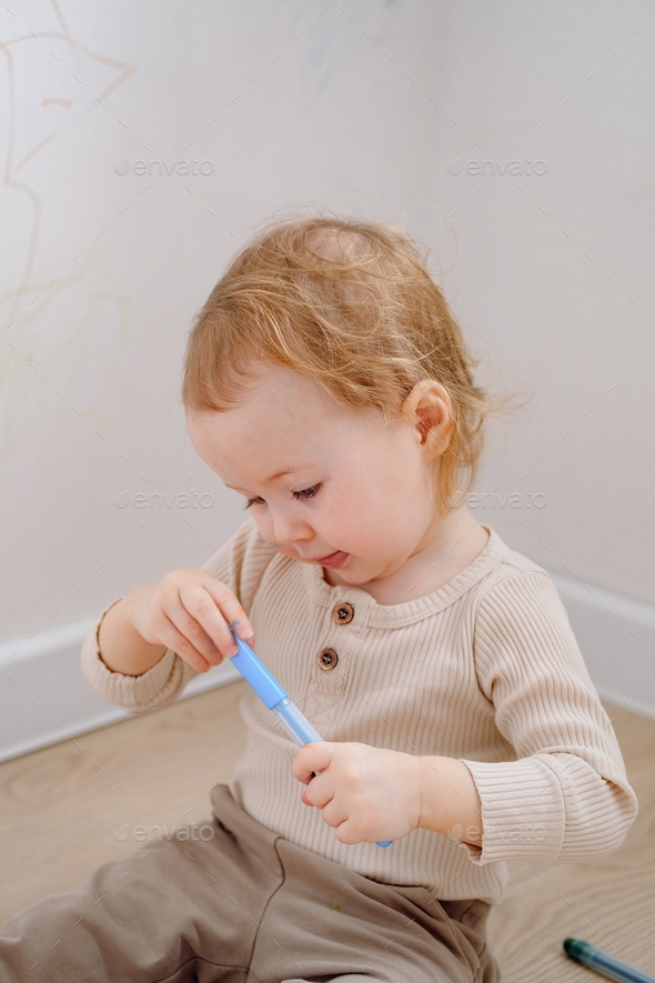 Close up of a cute toddler holding colored marker ready to draw on the white wall and himself.