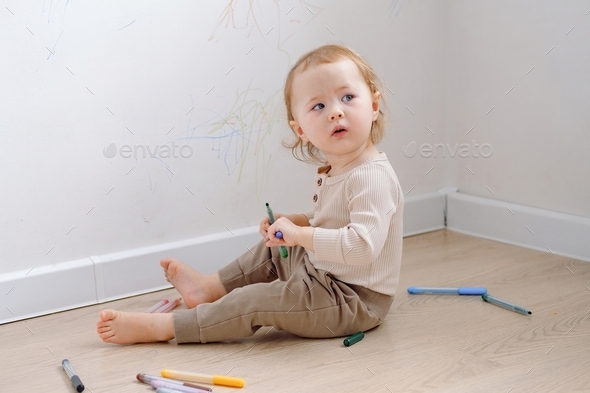 A toddler looking frightened sitting on the floor by white wall have been drawn by colored markers.