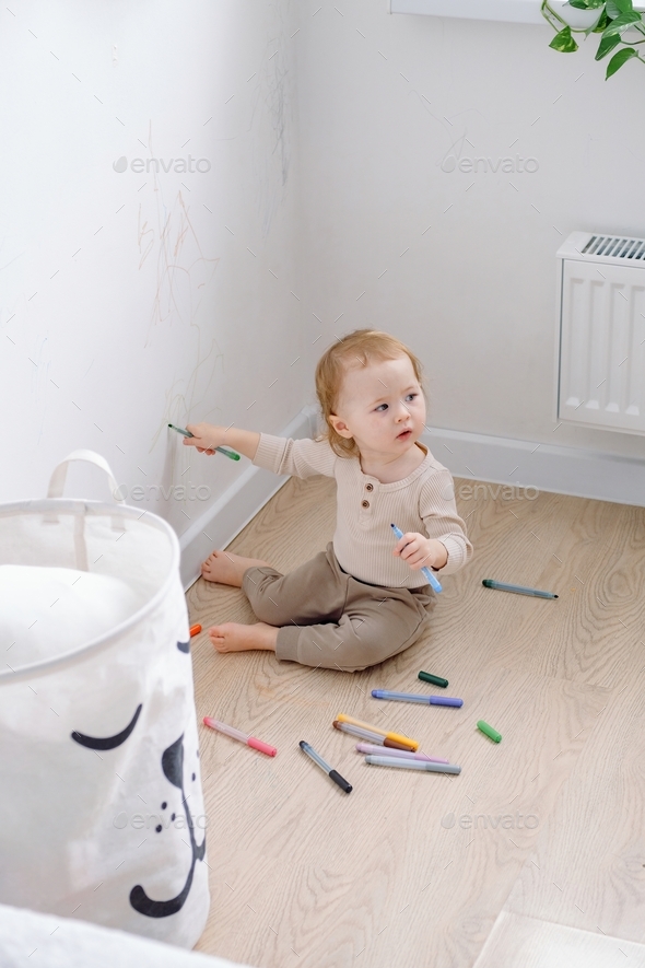 A toddler sitting by the white wall holding colored markers and drawing on the wall, looking away.
