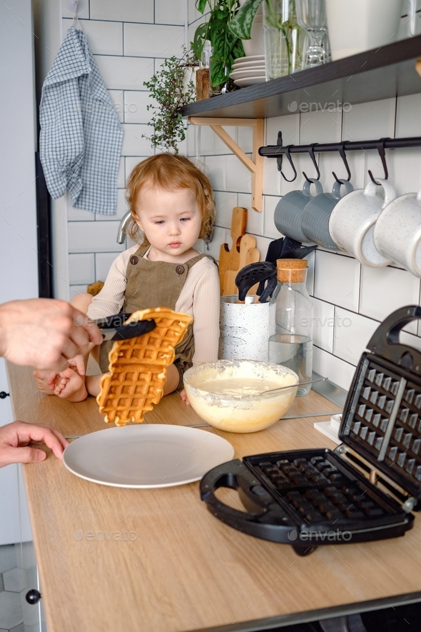 A toddler waiting for dad baking waffles for breakfast. A father and child spending time together