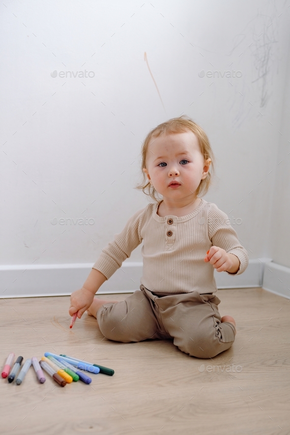 A toddler sitting by the white wall that has been drawn with colored markers and looking curiously.