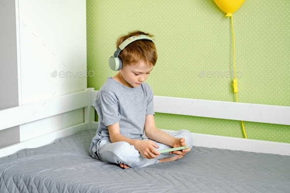 A preschool boy plays a game or watches a video on mobile using headphones. - Stock Photo - Images