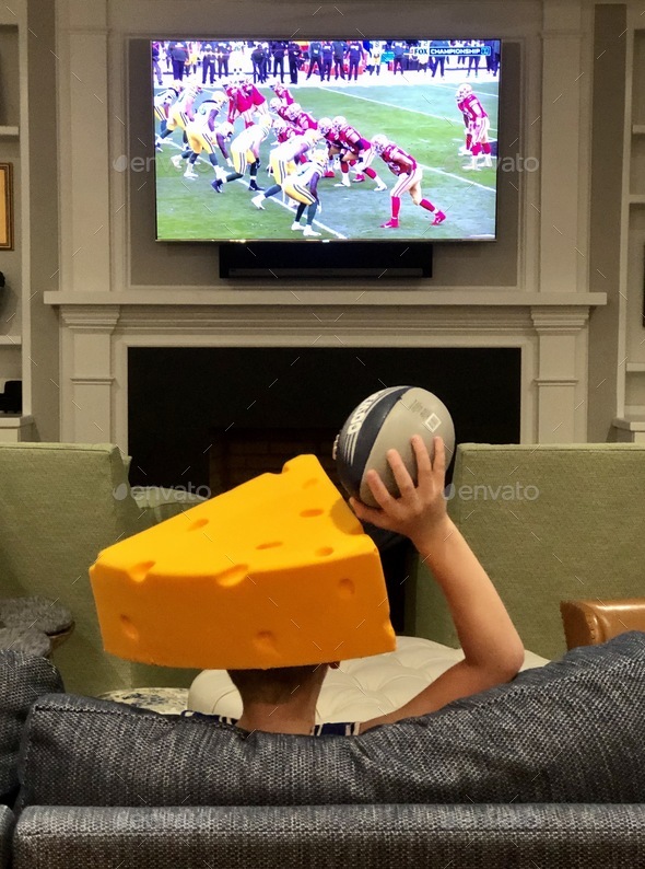 Child football fan watching football at home holding football wearing Green Bay packers cheese head - Stock Photo - Images