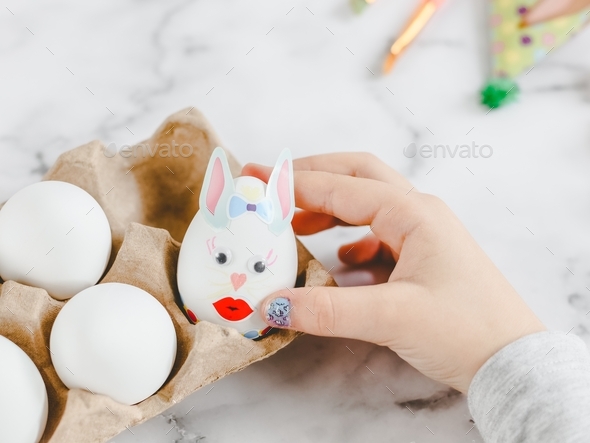 Hands of a caucasian girl in a gray turtleneck, laying an easter bunny egg with stickers in a cardbo