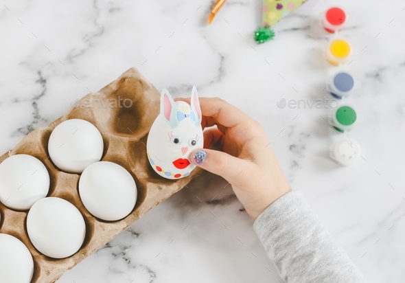 Hands of a caucasian girl in a gray turtleneck laying an Easter bunny egg with stickers in a cardboa