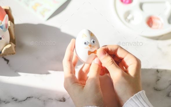 The hands of a caucasian girl in a white turtleneck glue an orange bow sticker on an easter bunny eg