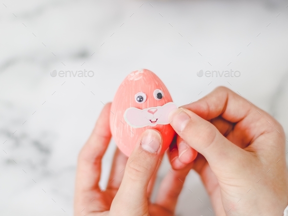 Hands of a caucasian girl pasting bow stickers on a pink acrylic painted easter egg with eyes