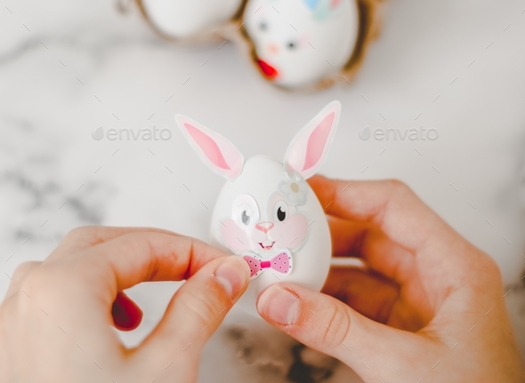 Hands of a caucasian girl pasting bow stickers on an easter bunny egg with ear