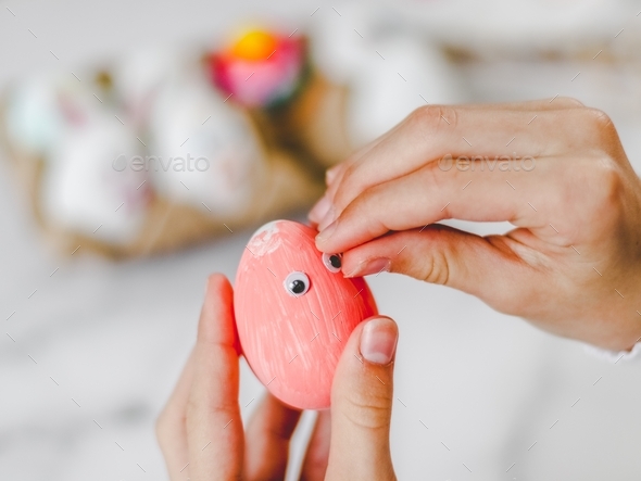 Hands of a caucasian girl sticking eye stickers on a pink acrylic painted easter egg