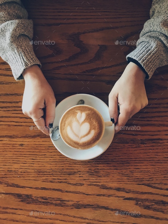 Beautiful cappuccino on a wooden table served hot and held by a woman’s hands  - Stock Photo - Images