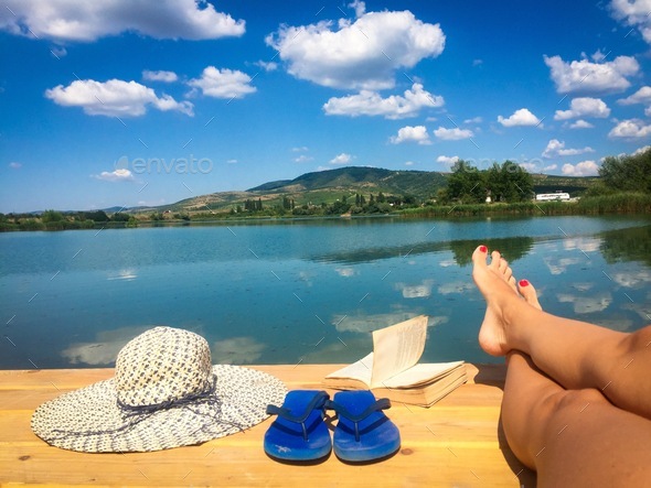 Summer hat,woman\'s feet, flip flops and a book on a wooden pontoon near the lake
