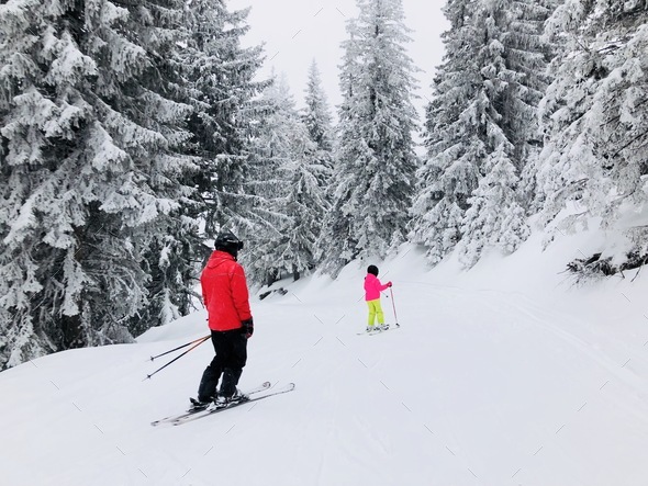 Adult skier in red jacket teaching little child to ski on a slope surrounded by conifers covered in