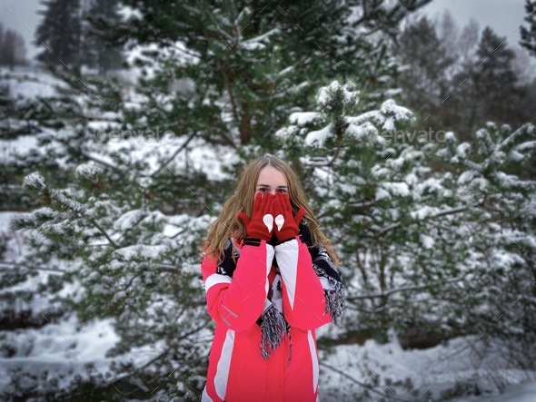 Portait of a woman in winter clothes wearing red gloves with hearts standing near a snowy pine tree
