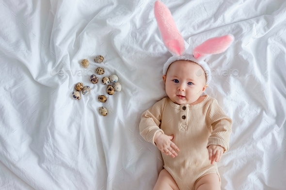 Cute funny baby with bunny ears dressed up for Easter laying on white bed sheet with quail eggs.  - Stock Photo - Images
