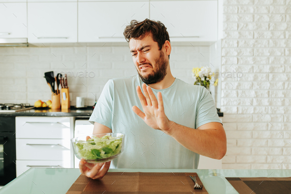 A young man, grimacing, looks towards the salad, how you don t want to eat it.
