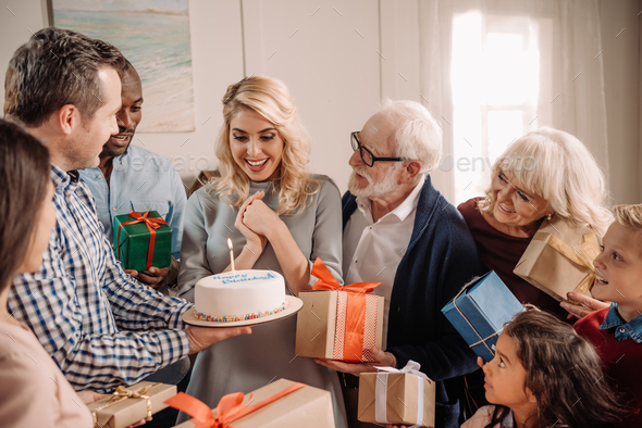 large family presenting cake and gifts to happy surprised woman