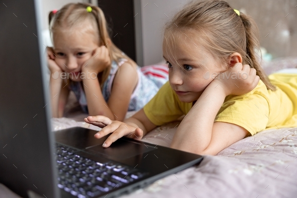Two sisters 7-8 years old playing a game on a laptop lying on the bed in the room. - Stock Photo - Images