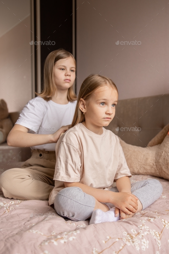 Two sisters are sitting on the bed in the room and the older sister is combing the younger one's hai - Stock Photo - Images