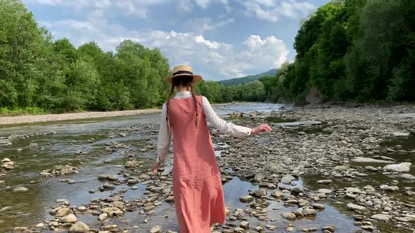 Vintage romantic woman in straw hat, long dress walks on river rocks near forest. Natural scenic