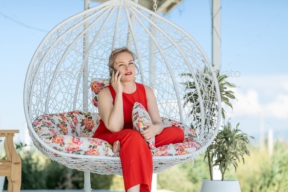 an attractive adult blonde plus size in a red jumpsuit is sitting in a cocoon hanging chair