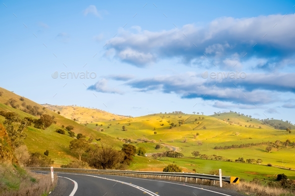 Open empty road surrounded by green hills, fields and farms in Australia. Road trip background