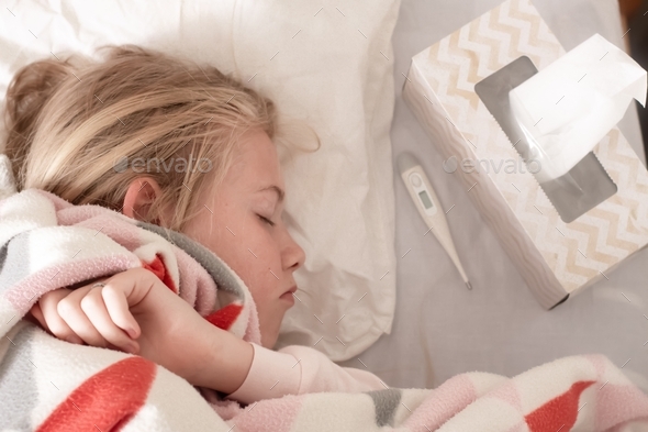 Sick teenage girl lying on the bed covered with blanket with thermometer