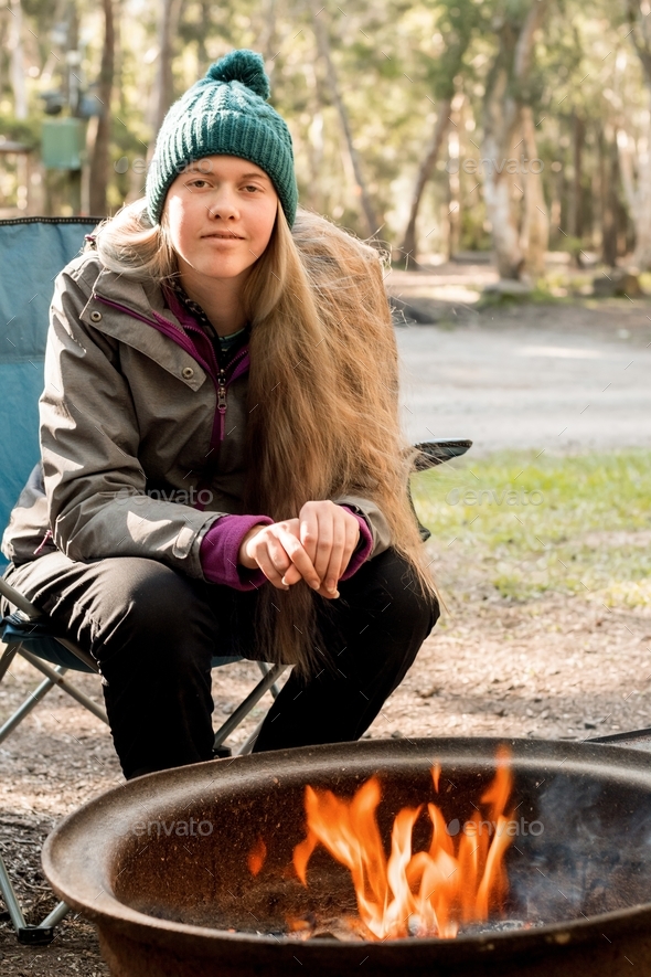 Teenage girl wearing coat and beanie sitting in a camping chair near campfire at the camping site