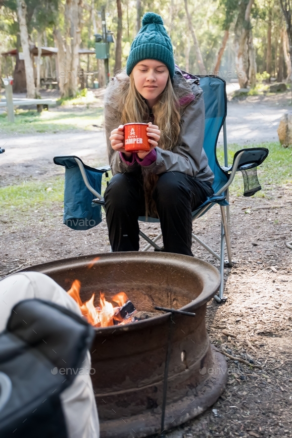Teenage girl wearing beanie sitting in a camping chair near campfire holding a red mug. Happy camper