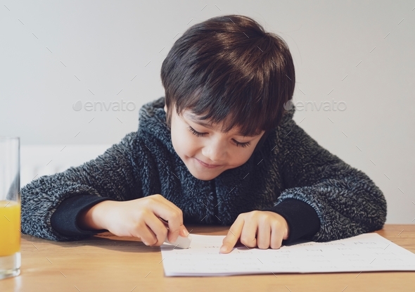 Kid doing homework with happy face. Cute child boy using rubber rubbing wrong words written on paper