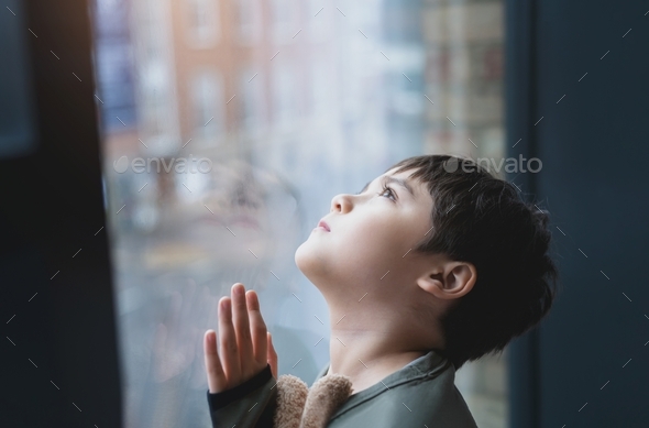 Kid standing next to window looking up to sky with curious face, Child looking through window glass