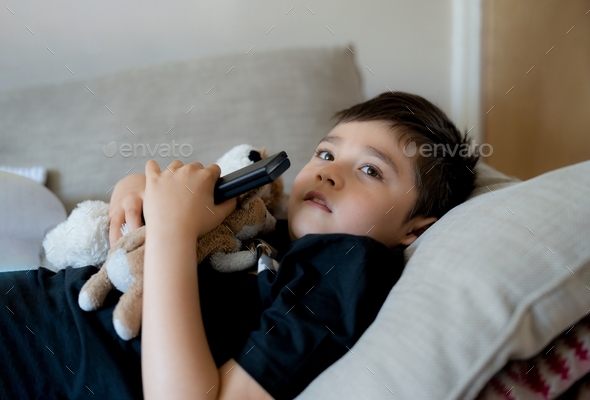 Boy holding remote control looking up with curious face,Kid sitting on sofa watching cartoon on TV