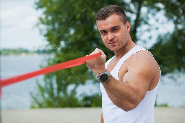 Athletic young man outdoors in summer, hard workout with elastic rubber loop expander.