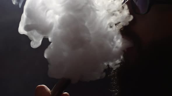 Young Bearded Man in Glasses Smokes a Hookah and Blow Out Smoke Closeup on Black Background in Slow