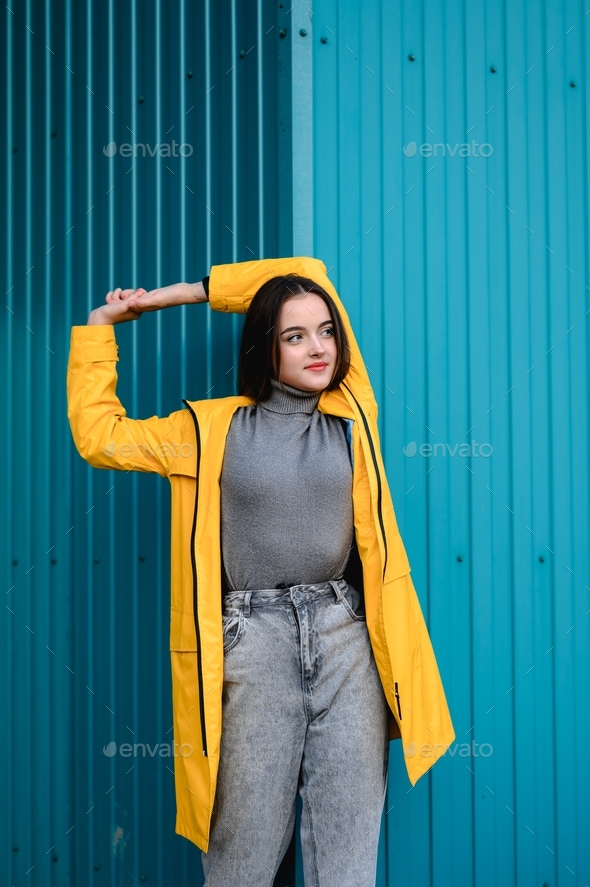 a young girl in a yellow raincoat and gray jeans stands near a blue wall.