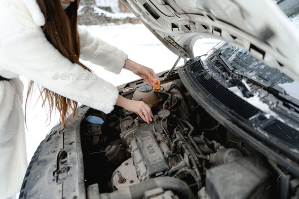on a road trip in winter, a girl broke her car. she has opened the hood and is trying to fix it.