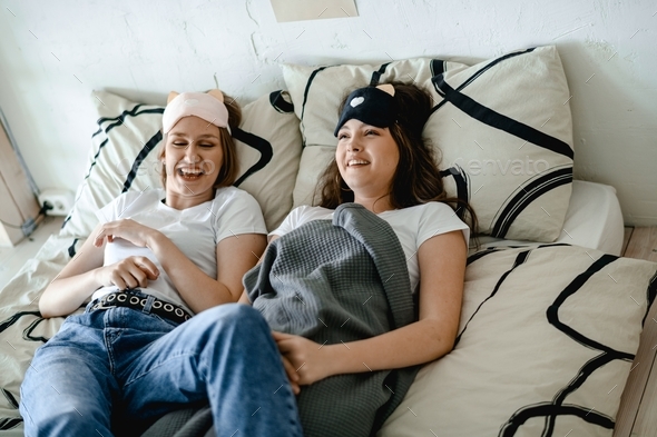 two girls in white T-shirts lie in bed. they wear sleep bands on their heads. They are laughing .