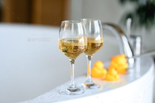 two glasses of champagne stand on the edge of a white tub. yellow rubber ducks stand behind them