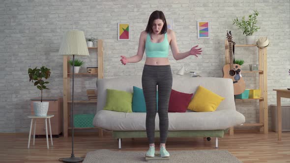 Expressive Distressed Young Woman in Sportswear Measures Her Weight on the Floor Scales alt