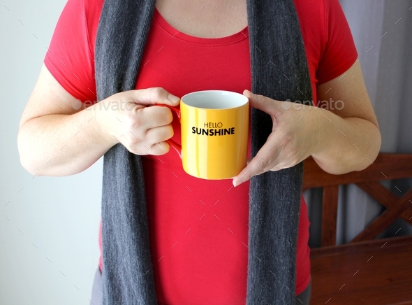 Middle aged woman holding a yellow cup of coffee in the morning that says Hello Sunshine on it