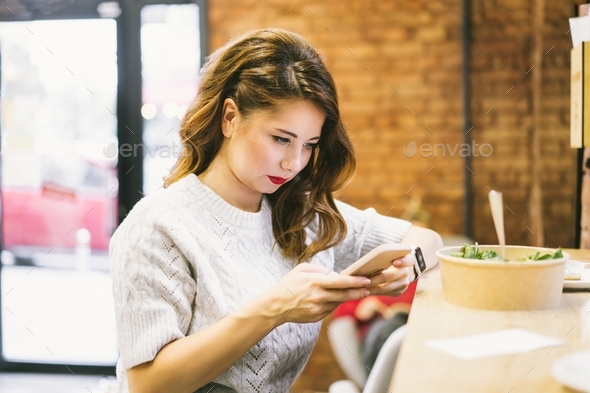 Concentrated woman looking on the phone while having lunch in the cafe. Remote work