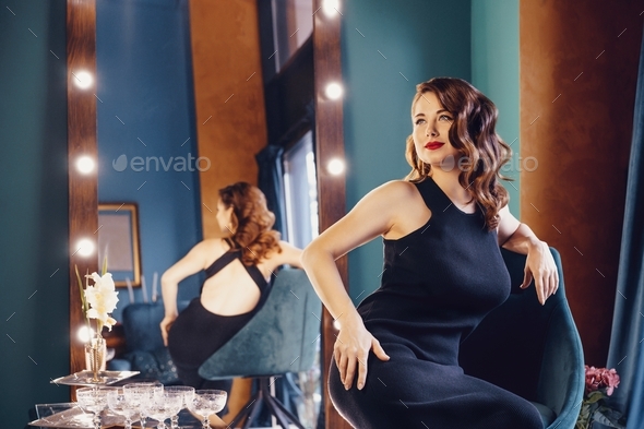 Posh woman with dark hair in elegant black dress posing over her reflection in illuminated mirror.