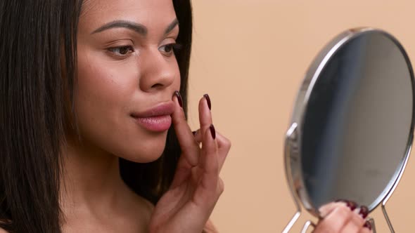 Black Woman Fixing Makeup Holding Mirror Over Beige Background SideView alt
