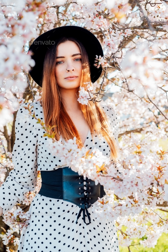 Young Brunette Woman In Hat And Dress Near A White Blooming Tree