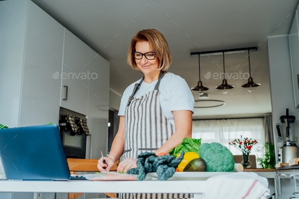 Middle age woman following a cooking tutorial video course on the laptop while preparing meal