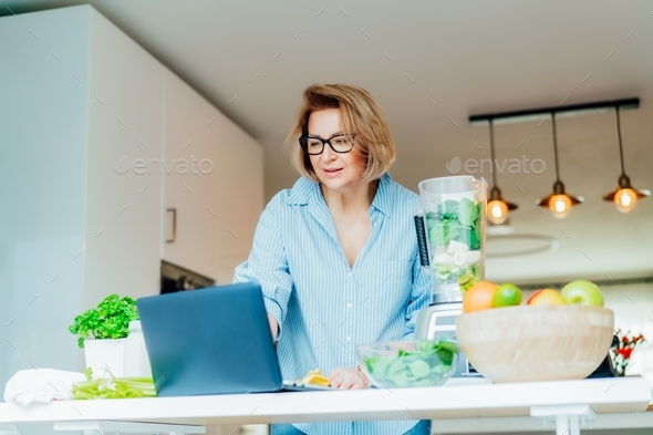 Woman following a cooking tutorial video course on laptop while preparing green smoothie in kitchen.