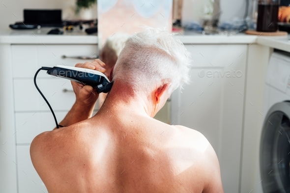 Back view white, silver hair man doing self haircut with a clipper and looks in the mirror.